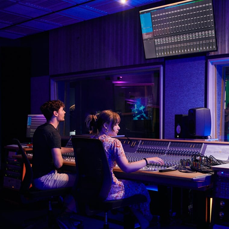 Two students sitting in a production booth, with performers in the background mobile