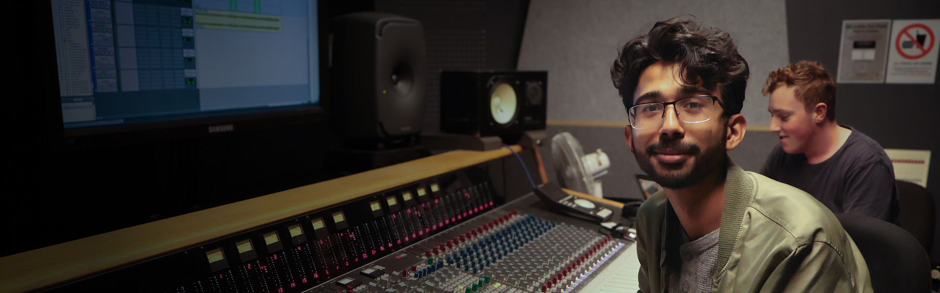 A young man with glasses sitting at a mixing desk and facing to camera - image for desktop