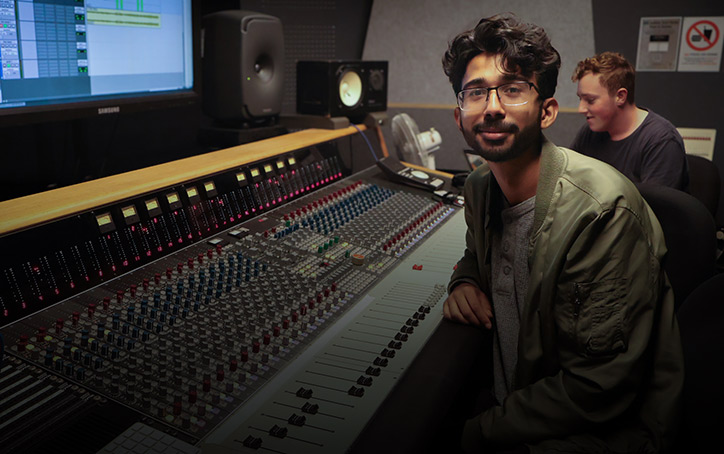 A young man with glasses sitting at a mixing desk and facing to camera - image for mobile