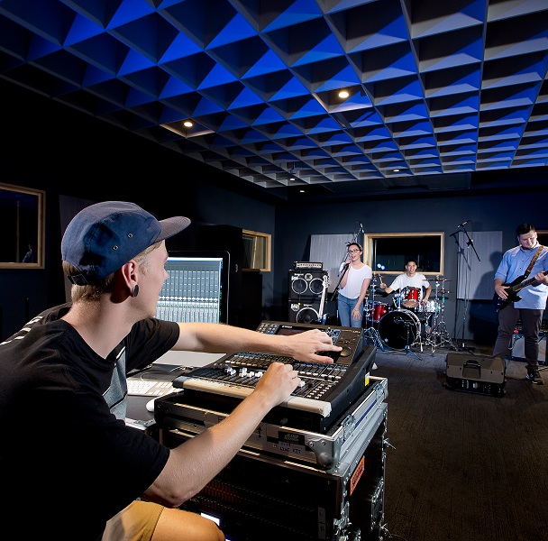 A man wearing a blue cap operating a channel mixer in a music studio, with a singer, guitar player and drummer playing in the background