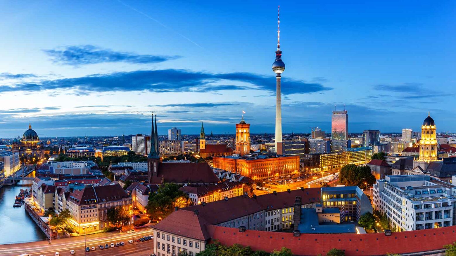 Cityscape of Berlin featuring the iconic Fernsehturm (TV Tower) rising above a skyline of historic and modern buildings under a clear sky.