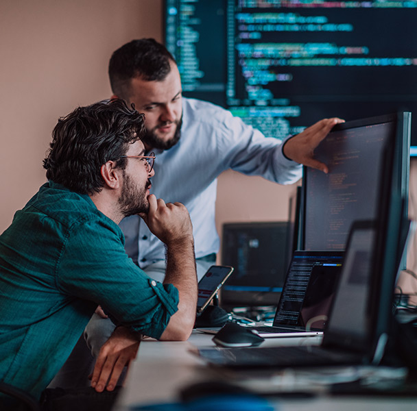 A male student with glasses sitting at a desk and looking towards a computer monitor with his hand on his chin, while a male teacher points towards the screen