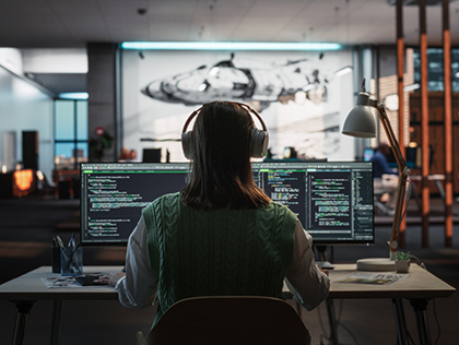 A women wearing a sweater and over ear headphones, working on two computer monitors - MIT scholarship tile image