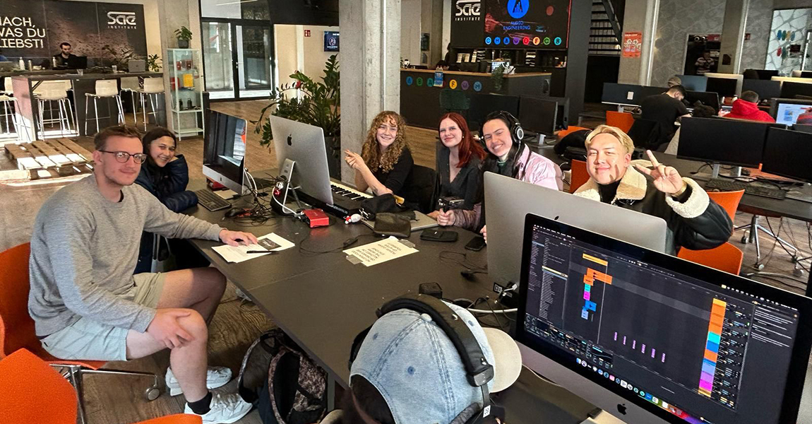 A group of six students sitting around a desk and working on desktop computers at an SAE campus