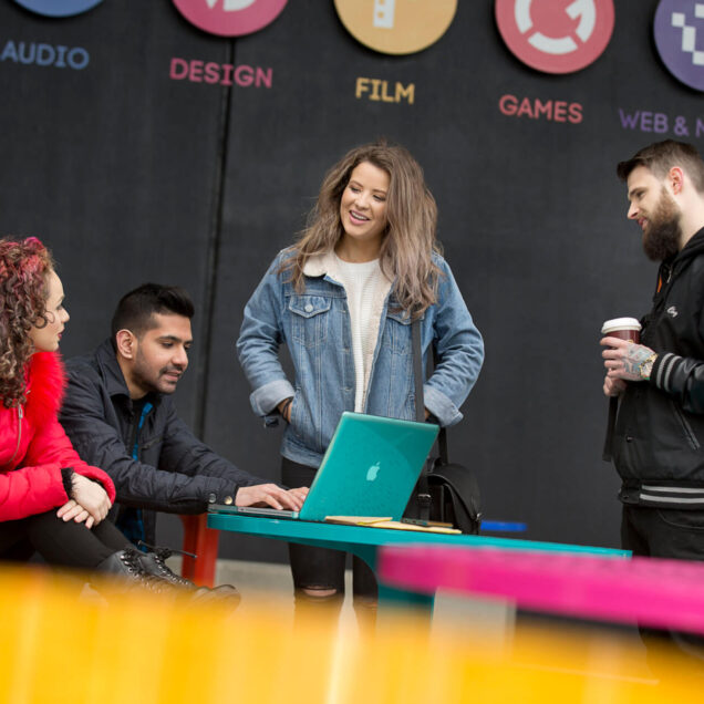 Melbourne Students stand in Front of Campus at desk. One at a laptop. One standing holds a coffee. One has has in pockets.