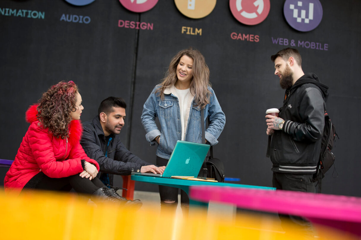 Melbourne Students stand in Front of Campus at desk. One at a laptop. One standing holds a coffee. One has has in pockets.