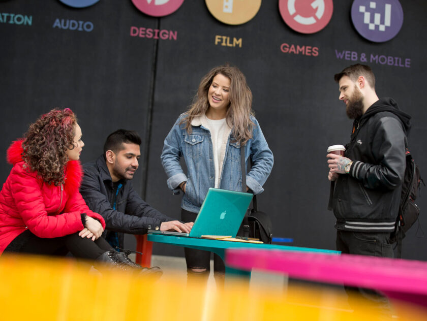 Melbourne Students stand in Front of Campus at desk. One at a laptop. One standing holds a coffee. One has has in pockets.