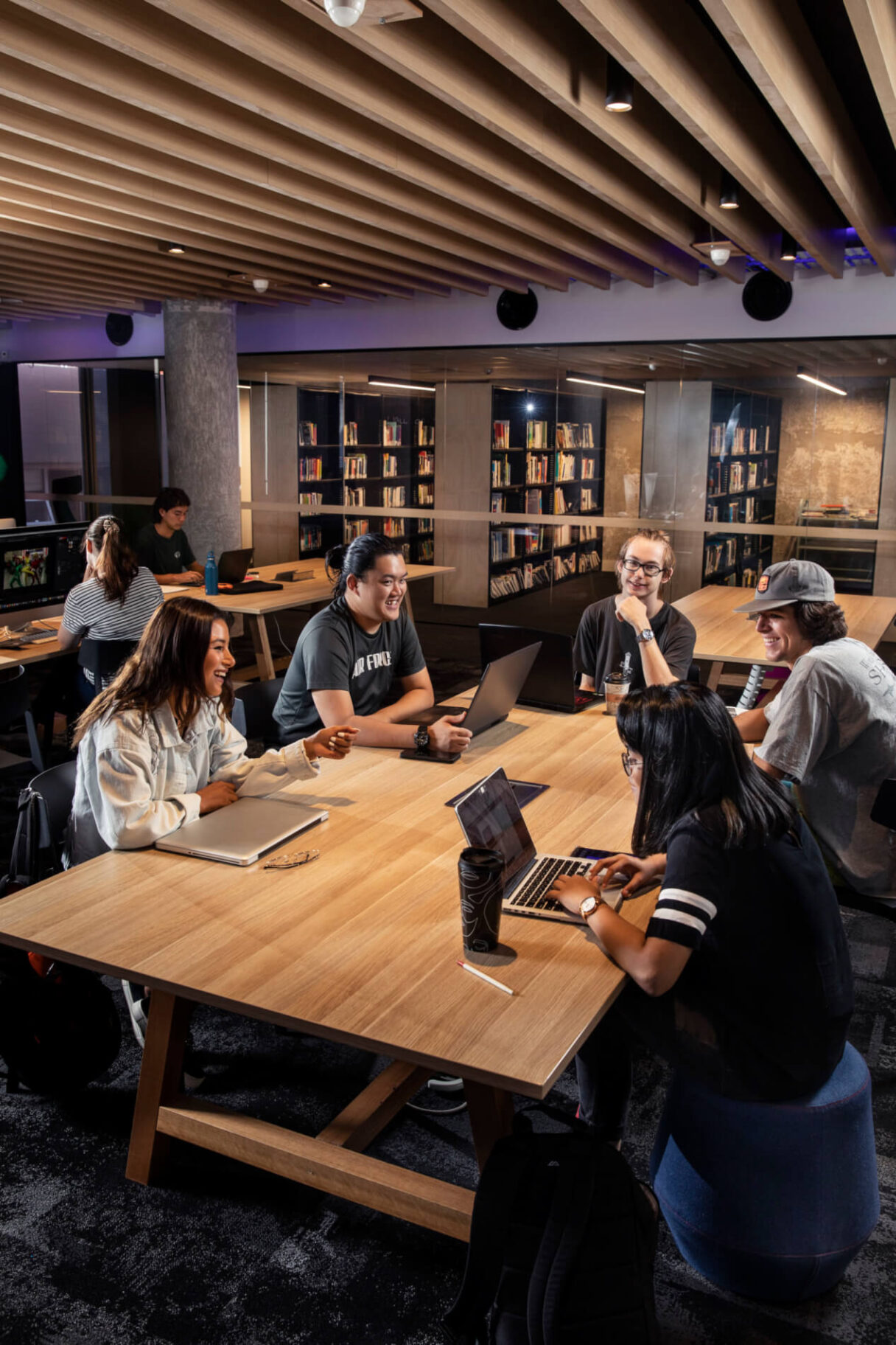 A group of students sit around a large wooden table with laptops. Laughing and looking at screen or each other.