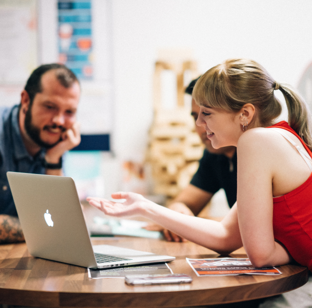 Three students sitting at a round desk, looking towards a MacBook screen