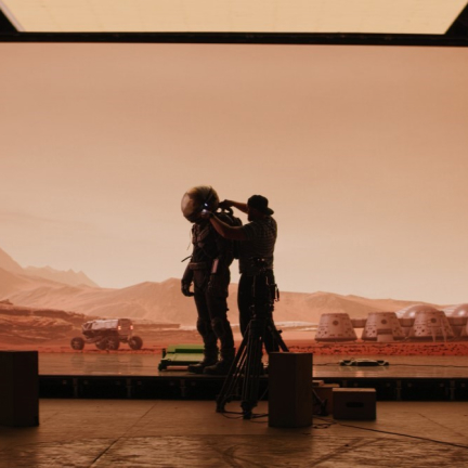 A producer and subject wearing an astronaut helmet, standing in front of a space backdrop