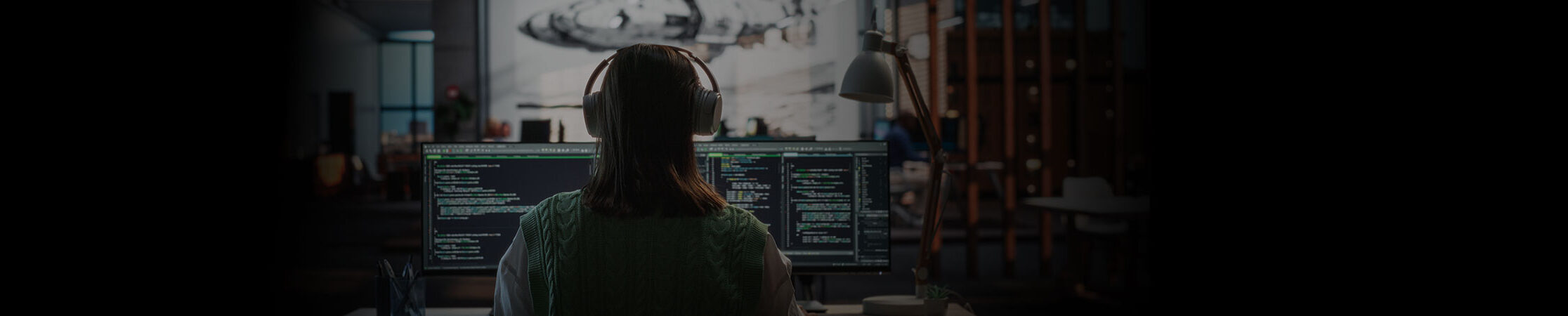 Person wearing wireless over ear headphones sits in officer - with two monitors in front.
