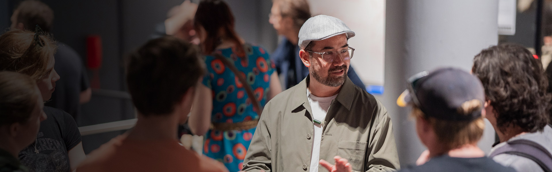 An SAE University College teacher wearing classes and a flat cap showing students around a college event - image for desktop