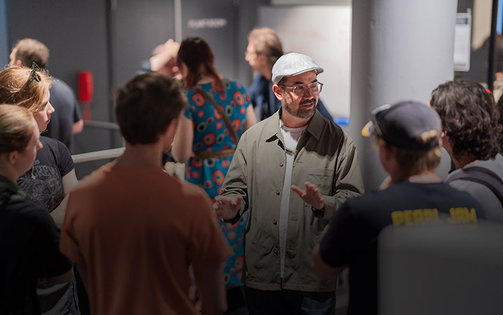 An SAE University College teacher wearing classes and a flat cap showing students around a college event - image for mobile