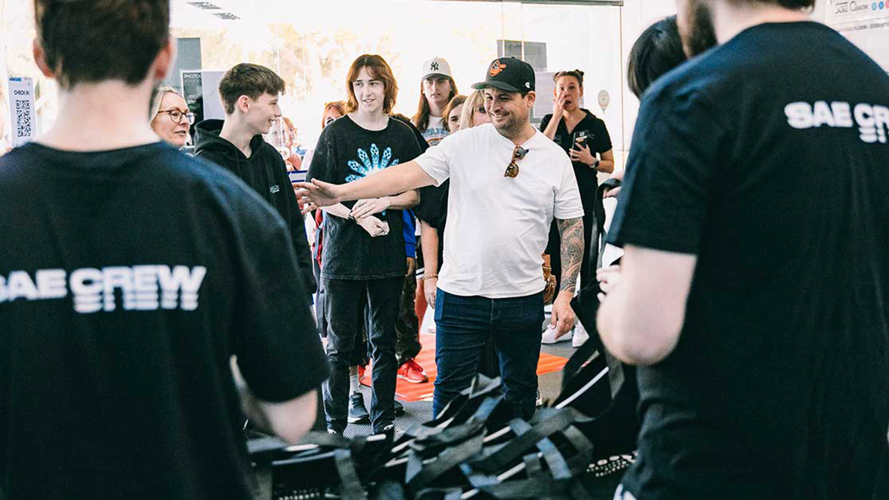 A male teacher from SAE University College standing at an Info Night event, surrounded by SAE crew and event attendees