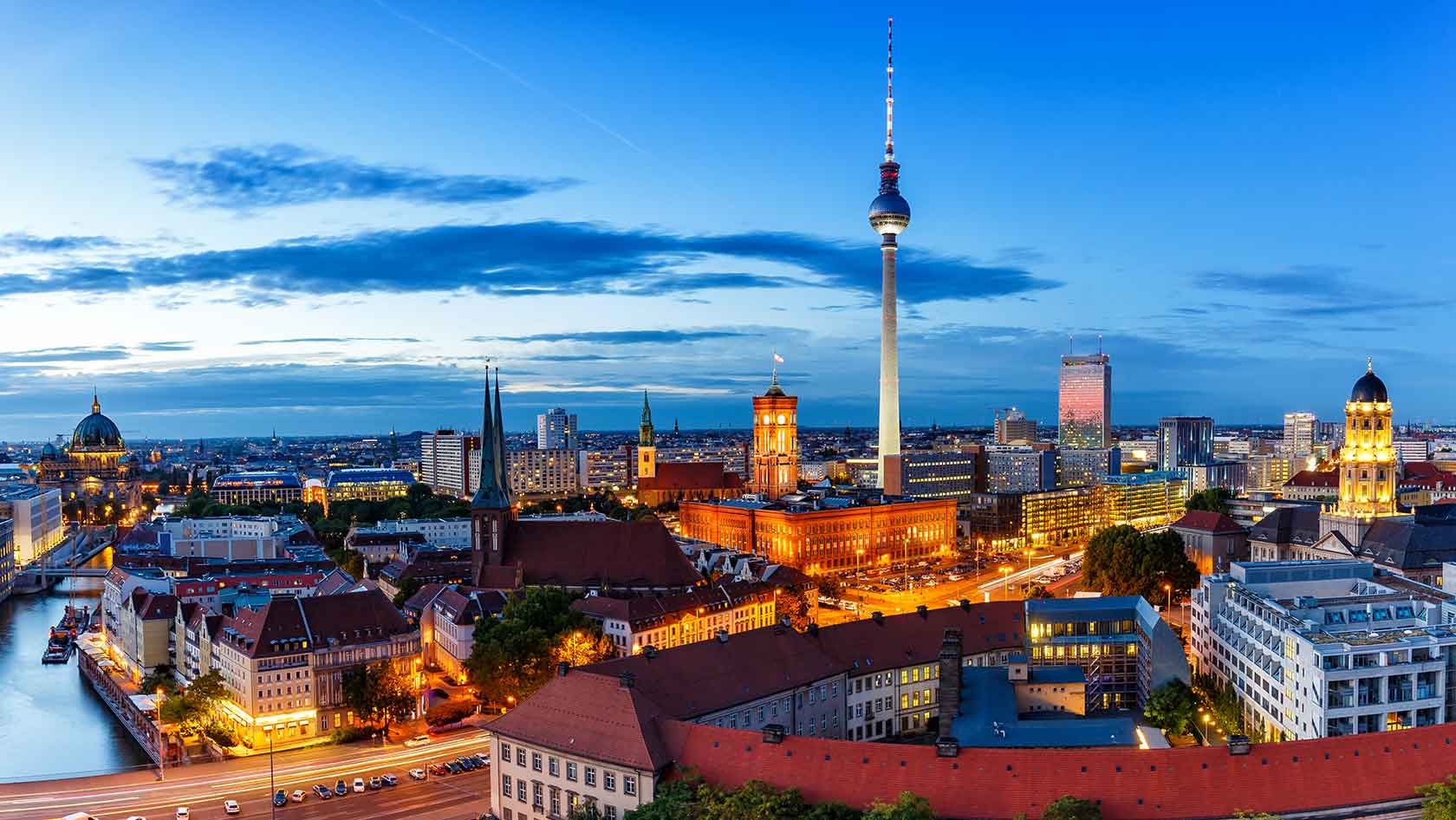 Cityscape of Berlin featuring the iconic Fernsehturm (TV Tower) rising above a skyline of historic and modern buildings under a clear sky.