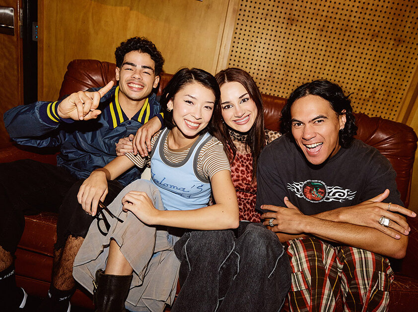 Four students sitting together on a red leather couch in a studio with wooden walls, smiling toward camera - SAE Open House event Sydney (1116x628)