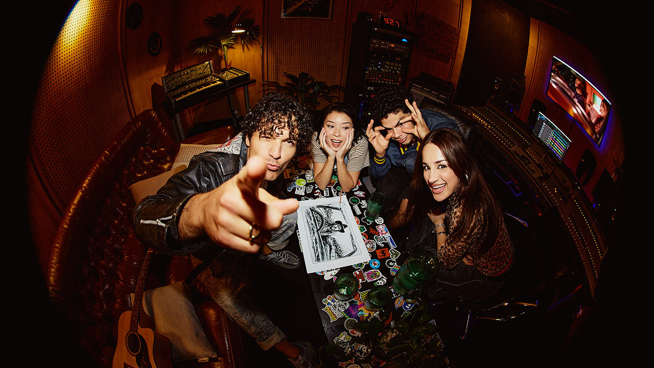Four SAE University College students shot with a fish eye lens - standing around a desk covered in stickers, with one pointing upwards towards camera - SAE Open House event (1280x720)