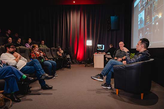 a man and woman in armchairs on stage with microphone. the woman is speaking and man is looking at a laptop